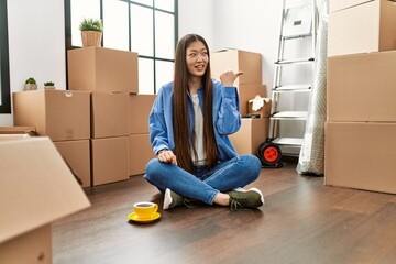 Wall Mural - Young chinese girl sitting on the floor at new home smiling with happy face looking and pointing to the side with thumb up.