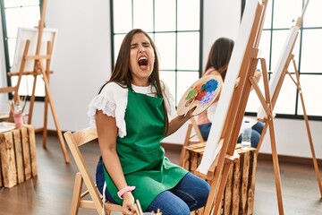 Wall Mural - Young hispanic artist women painting on canvas at art studio angry and mad screaming frustrated and furious, shouting with anger. rage and aggressive concept.