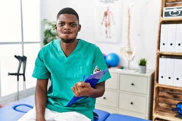 Sticker - Young african american man working at pain recovery clinic puffing cheeks with funny face. mouth inflated with air, crazy expression.