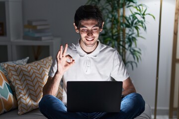 Poster - Young hispanic man using laptop at home at night smiling positive doing ok sign with hand and fingers. successful expression.