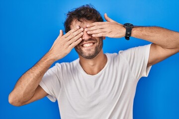 Poster - Hispanic young man standing over blue background covering eyes with hands smiling cheerful and funny. blind concept.