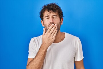 Sticker - Hispanic young man standing over blue background bored yawning tired covering mouth with hand. restless and sleepiness.