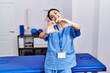 © Krakenimages.com - Young hispanic woman wearing physiotherapist uniform standing at clinic smiling in love doing heart symbol shape with hands. romantic concept.