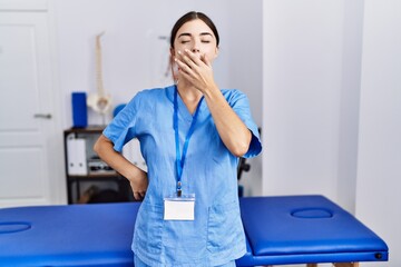 Canvas Print - Young hispanic woman wearing physiotherapist uniform standing at clinic bored yawning tired covering mouth with hand. restless and sleepiness.