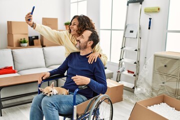 Poster - Middle age hispanic couple smiling happy. Man sitting on wheelchair with dog on his legs and woman making selfie by the smartphone at new home.