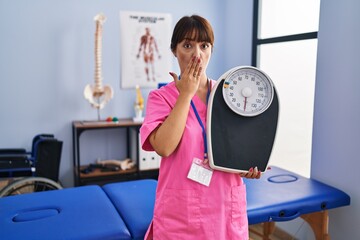 Canvas Print - Young brunette woman as nutritionist holding weighing machine covering mouth with hand, shocked and afraid for mistake. surprised expression