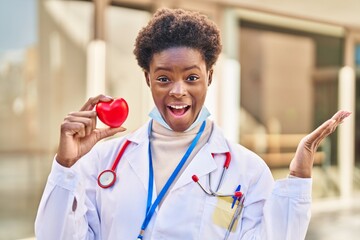 Sticker - African american woman wearing doctor uniform holding heart celebrating victory with happy smile and winner expression with raised hands