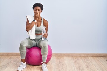 Wall Mural - African american woman wearing sportswear sitting on pilates ball laughing and embarrassed giggle covering mouth with hands, gossip and scandal concept