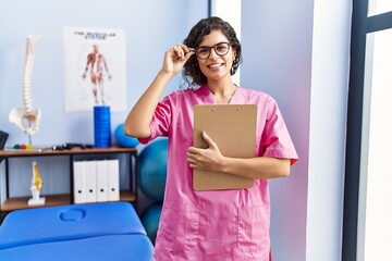 Poster - Young latin woman wearing physiotherapist uniform holding clipboard at clinic