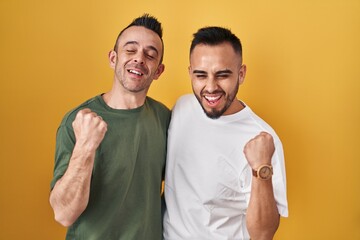 Poster - Homosexual couple standing over yellow background very happy and excited doing winner gesture with arms raised, smiling and screaming for success. celebration concept.