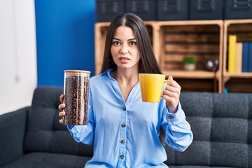 Poster - Young brunette woman holding coffee beans and cup of coffee clueless and confused expression. doubt concept.