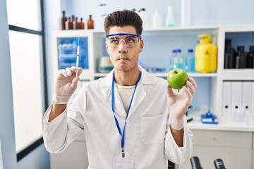 Wall Mural - Young hispanic man working at scientist laboratory holding apple making fish face with mouth and squinting eyes, crazy and comical.