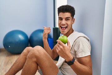 Sticker - Young hispanic man wearing sportswear eating healthy apple pointing thumb up to the side smiling happy with open mouth