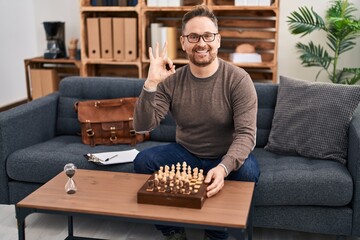 Sticker - Middle age caucasian man playing chess sitting on the sofa doing ok sign with fingers, smiling friendly gesturing excellent symbol