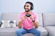 © Krakenimages.com - Hispanic young man playing video game holding controller sitting on the sofa looking away to side with smile on face, natural expression. laughing confident.