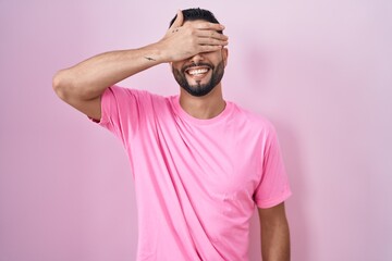 Poster - Hispanic young man standing over pink background smiling and laughing with hand on face covering eyes for surprise. blind concept.