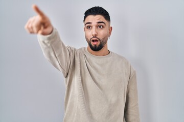 Poster - Young handsome man standing over isolated background pointing with finger surprised ahead, open mouth amazed expression, something on the front