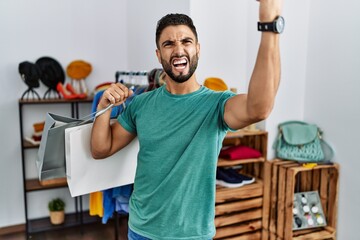 Wall Mural - Young handsome man with beard holding shopping bags at retail shop angry and mad raising fist frustrated and furious while shouting with anger. rage and aggressive concept.