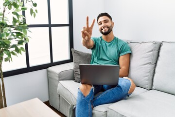 Sticker - Young handsome man with beard using computer laptop sitting on the sofa at home smiling looking to the camera showing fingers doing victory sign. number two.