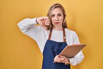 Sticker - Young blonde woman wearing professional waitress apron holding clipboard with angry face, negative sign showing dislike with thumbs down, rejection concept