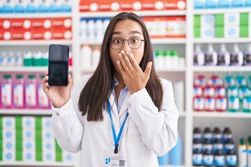 Sticker - Young hispanic woman working at pharmacy drugstore showing smartphone screen covering mouth with hand, shocked and afraid for mistake. surprised expression