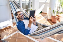Middle Age Man Reading Book Lying On Hammock At Terrace Home