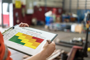 Wall Mural - a manager is holding a pen and risk matrix form with factory workshop place as blurred background. i