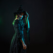 © Ulia Koltyrina - A charming young witch in a costume invites you to a sabbath, a Halloween party with costumes. Young brunette in pointed hat and black dress, photo on black