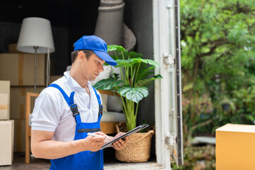 Wall Mural - Man mover worker in blue uniform checking lists on clipboard while unloading cardboard boxes from truck.Professional delivery and moving service.