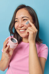 Wall Mural - Skin hydration concept. Positive lovely Asian woman applies moisturising cream on face smiles gladfully dressed in casual pink t shirt enjoys cosmetic procedures isolated over blue background