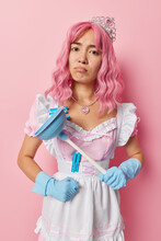 Vertical Shot Of Displeased Of Young Asian Housemaid With Pink Hair Holds Plunger Washes Toilet Wears Crown And Dress Does Domestic Chores Looks Sadly At Camera Isolated Over Rosy Background