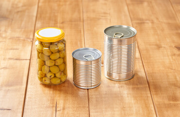 Iron tin can with tab opener and olives in a glass jar on the wooden table.