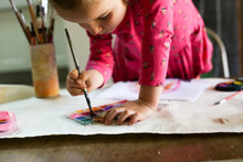 Girl Wearing Pink Dress Holding A Paintbrush And Dipping It In Water Colour Paint