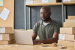© Mediaphotos - African worker typing on laptop making delivery online while sitting at table with stack of parcels