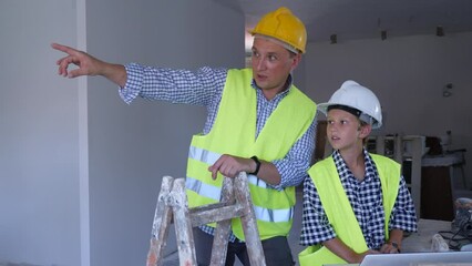 Wall Mural - Young boy and his father wearing protective helmets and yellow vests, discussing work plan in new apartment, using laptop. Man making pointing finger gesture.