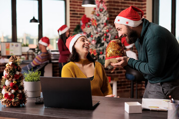 Businessman giving present box to woman in office, celebrating christmas eve holiday with gifts. Coworkers feeling happy about festive season with xmas ornaments and decorations.