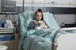 Ill kid resting in children healthcare facility patient bed alone while having teddy bear. Sick little girl under treatment wearing oxygen tube while holding plush bear toy.