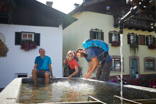 Happy Hiker Sitting By Playful Friends Splashing Water At Fountain In Village, Mutters, Tyrol, Austria