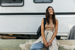 Pretty young caucasian woman enjoys spending time sitting by white motorhome. Brunette looks away, wears tank top, shirt, backpack and jeans. Relaxation concept