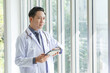 © aekkorn - Portrait of asian senior doctor standing and checking time in medical office while using tablet.