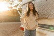 Cheerful young caucasian brunette woman posing looking at camera standing near auto house at sunset. Girl is wearing beige hoodie and jeans. Rest time concept.