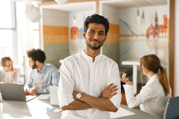 portrait of smiling indian businessman standing in modern office on colleagues background