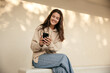 Positive young caucasian woman uses modern smartphone sitting against white wall. Brunette smiles, wears hoodie and jeans. Social media addiction concept