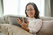 © bongkarn - Happy Asian-aged woman in eyeglasses sits on sofa in living room, using digital tablet.