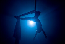 A Young Woman Performing In A Circus On Aerial Silk In The Dark With Blue Light. Silhouette Of A Female Equilibrist Hanging Upside Down And Demonstrating The Twine. Performance Of Aerial Gymnast.