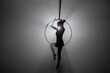 Silhouette of flexible aerial gymnast on an aerial ring performs acrobatic tricks in the air. Young woman performs with circus show in dark smoky studio with backlight.