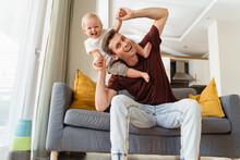 Father And Son Relationships. Handsome Dad Carrying Baby Boy On His Shoulders, Bending To Side Pretending To Fall Making Cute Kid Laugh With Excitement Sitting On Grey Sofa. Fatherhood, Parenting