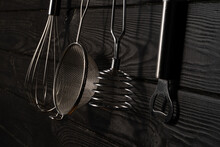 Opener, Whisk, Masher And Strainer Hanging Against Grey Wall In A Restaurant Or Home Kitchen. Kitchen Utensils Made Of Stainless Steel Or Metal. Set Of Tools For Cooking Food. Kitchenware On Drainer.