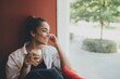 © polinaloves - Young woman drinking coffee cappuccino sitting near window.