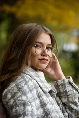 Poster - Close-up portrait of young woman in a stylish trench coat and in a white sweater resting on the bench. warm autumn day.